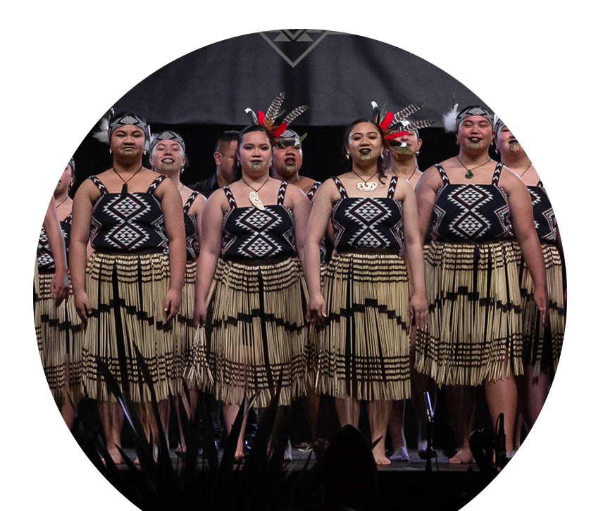 A kapa haka rōpū performs on stage, proudly adorned in black, white, and red kākahu - including traditional piu piu and a feathered Tīpare.