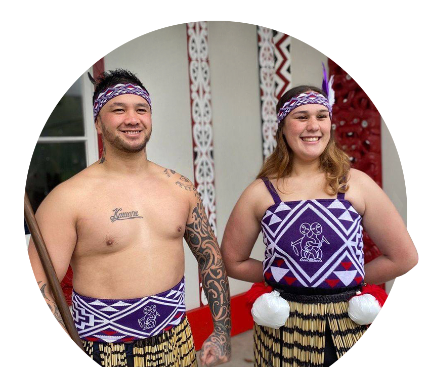 A proud Tāne and Wahine standing in front of a marae, dressed in custom-knitted purple, white and red Kākahu -made by Kapa Haka Makers.