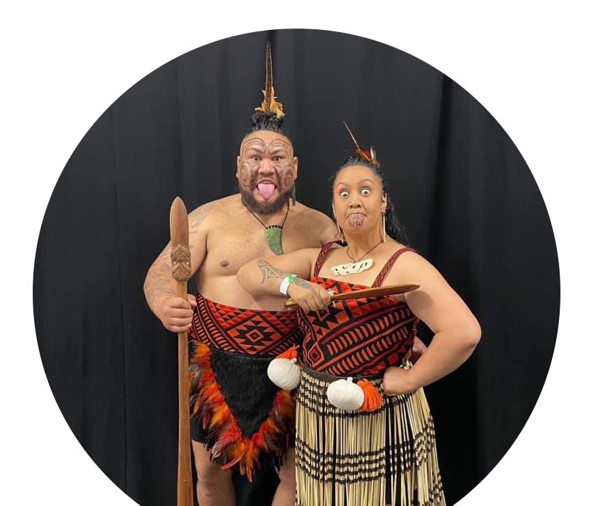 A Tāne and Wahine, dressed in traditional Māori kākahu in bold red and black, channel strength and intensity through a powerful pūkana.